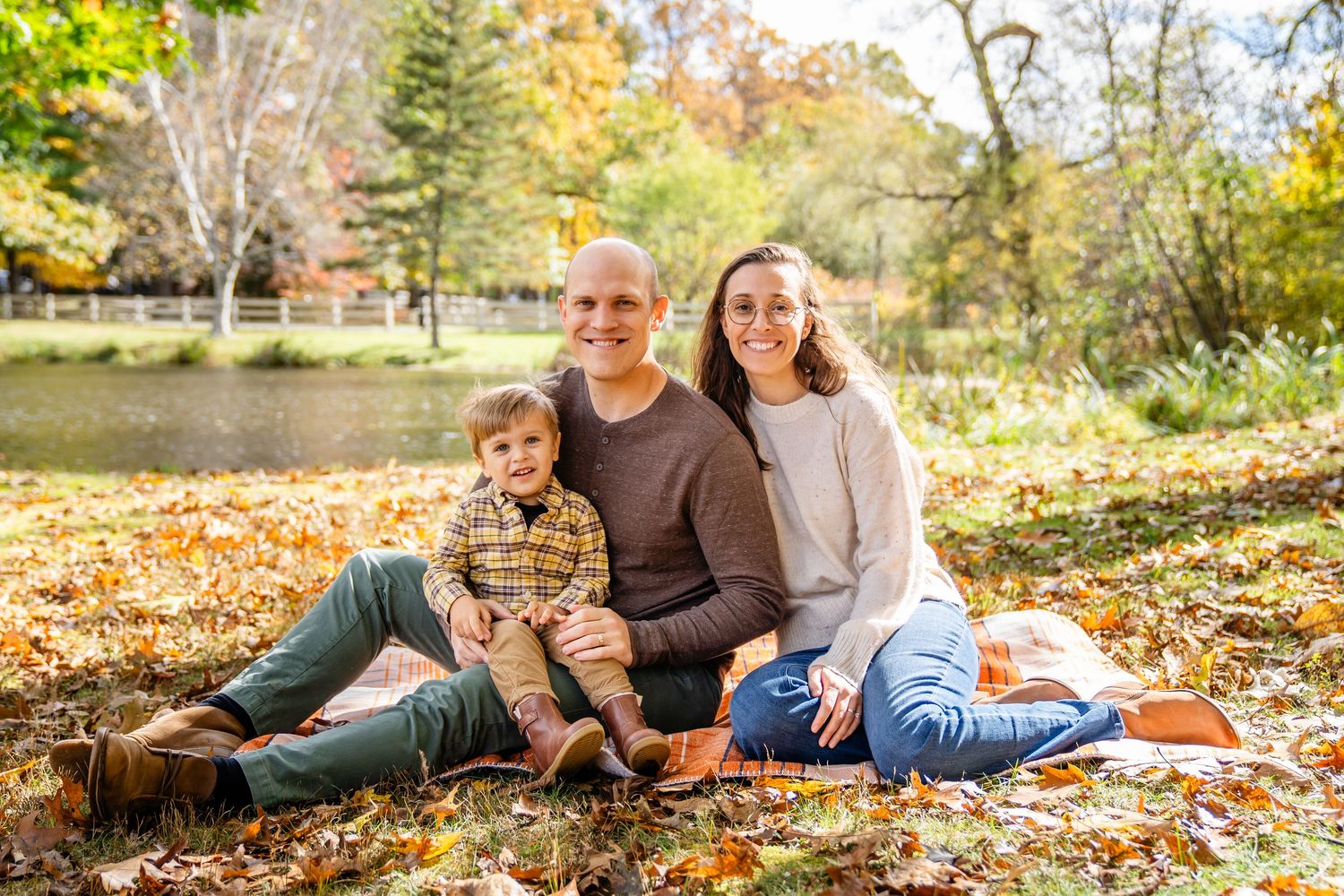 A family sits together on fallen autumn leaves near a pond on a sunny fall day in the park. North Shore, MA mini sessions. Endicott Park