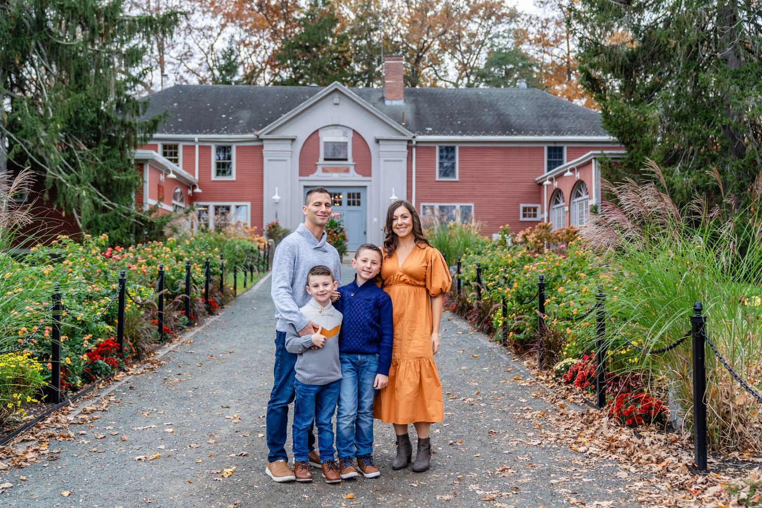 Family walks together on a fall path lined with autumn foliage in front of a red brick house. North Shore, MA mini sessions. Family Sessions
