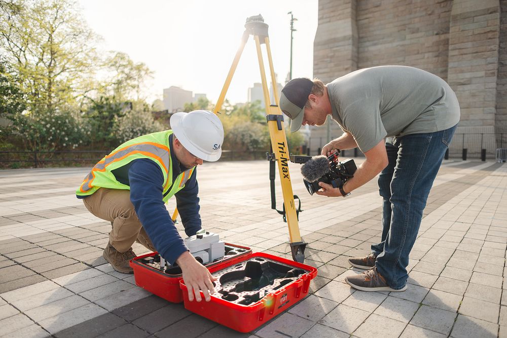 Land surveyors set up measuring equipment with a yellow theodolite and red tool case on a paved surface.