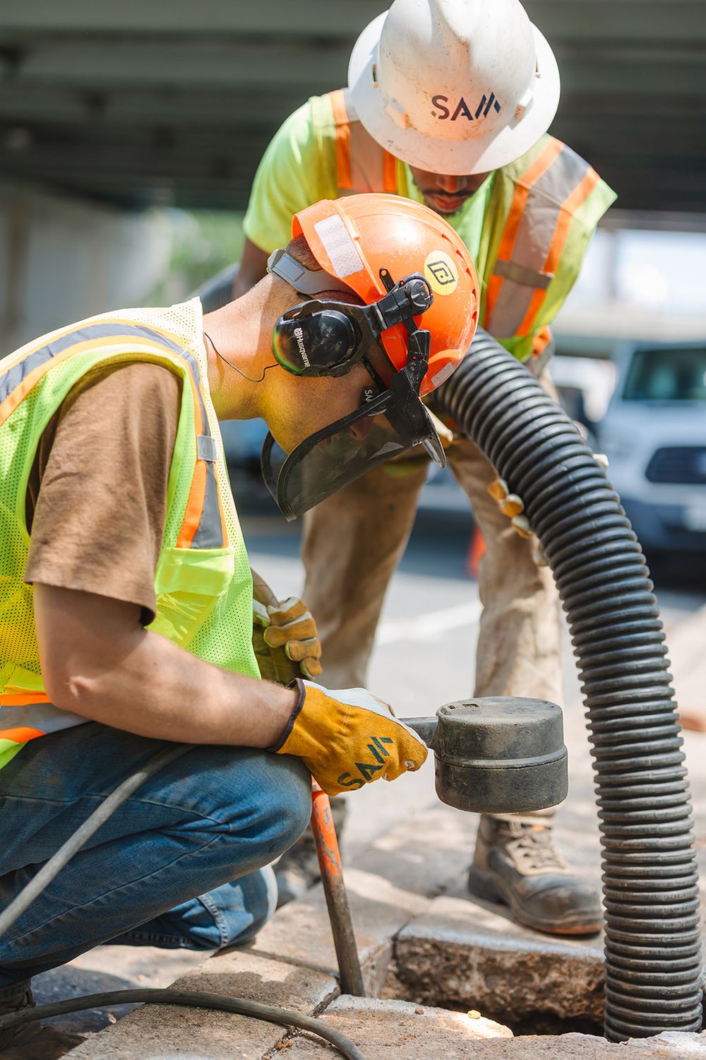 Construction workers in safety gear operate underground utility equipment and vacuum hose at a worksite.