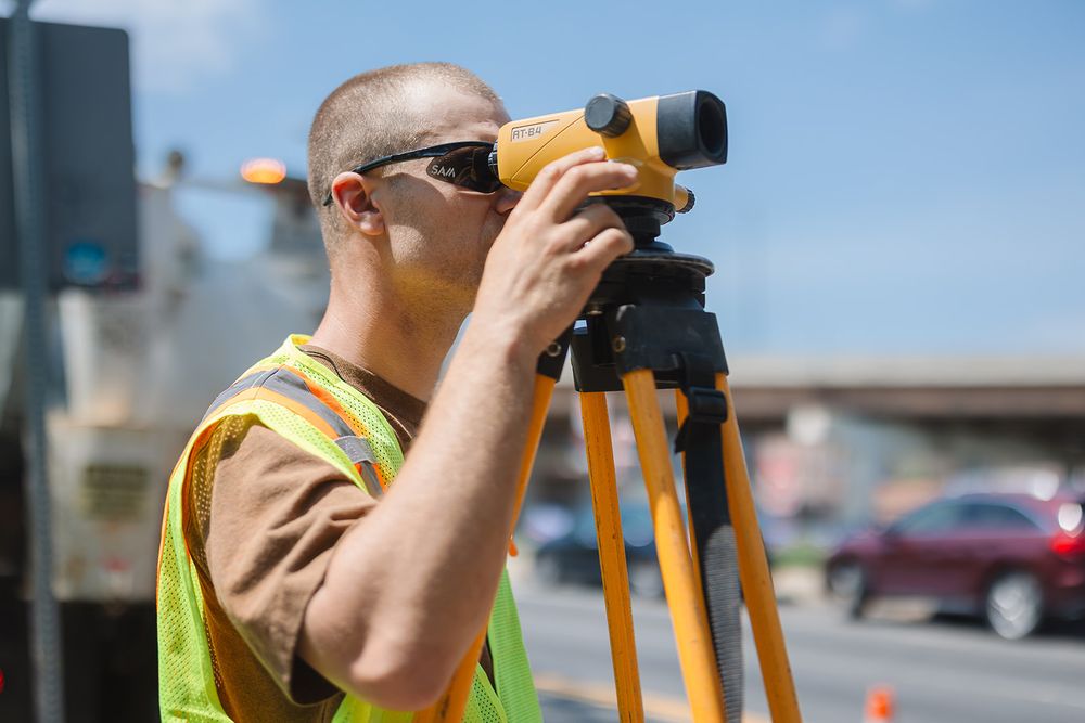 A surveyor in a high visibility vest operates a theodolite instrument on a construction site during daylight hours.