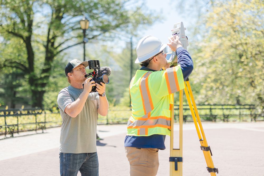 Construction surveyor using equipment on tripod while being photographed outdoors in park setting.