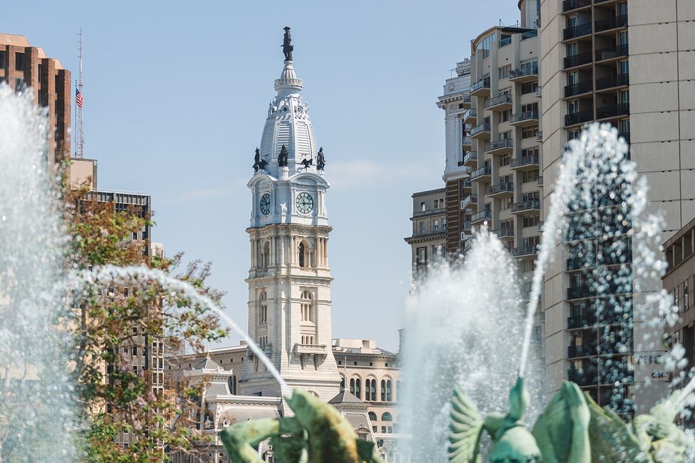 Philadelphia City Hall's ornate clock tower rises behind spouting fountain waters in Logan Square on a sunny day.