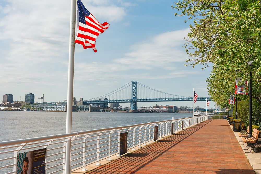 American flag waves along the Delaware River waterfront with Benjamin Franklin Bridge and Philadelphia skyline view.