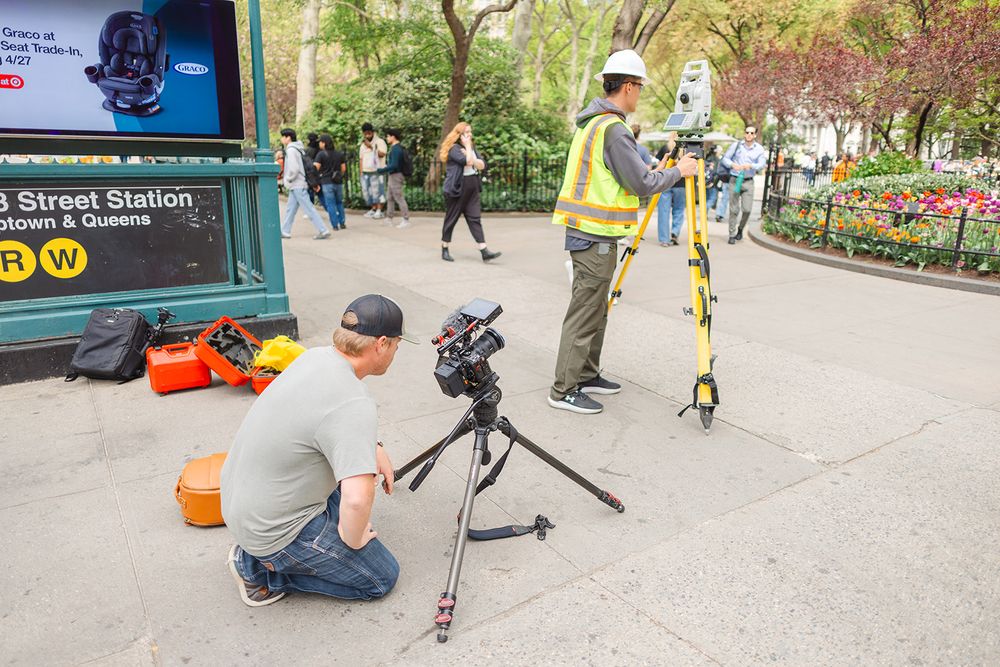 A camera crew sets up filming equipment near a subway station entrance with orange pumpkins on display.