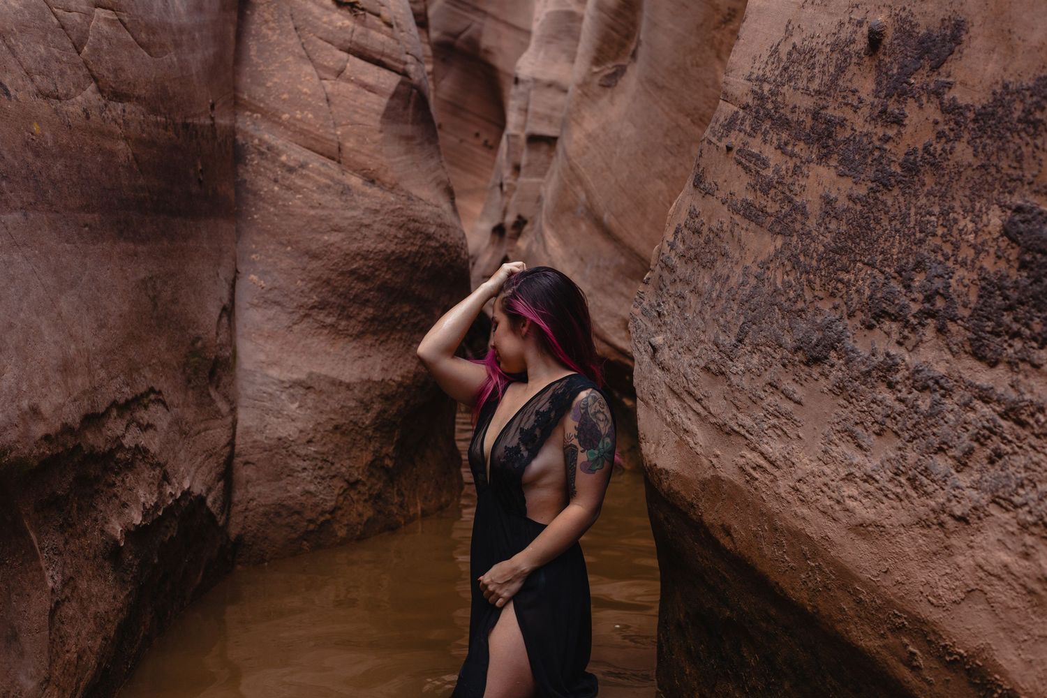 a tattoo woman with pink hair, posing casually between a flooded slot canyon, in a black lace dress