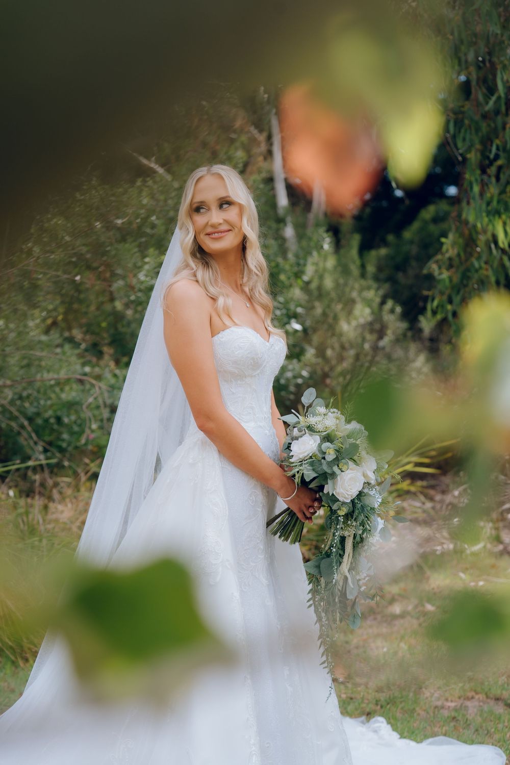 Bride in white lace gown poses with bouquet amid garden greenery.