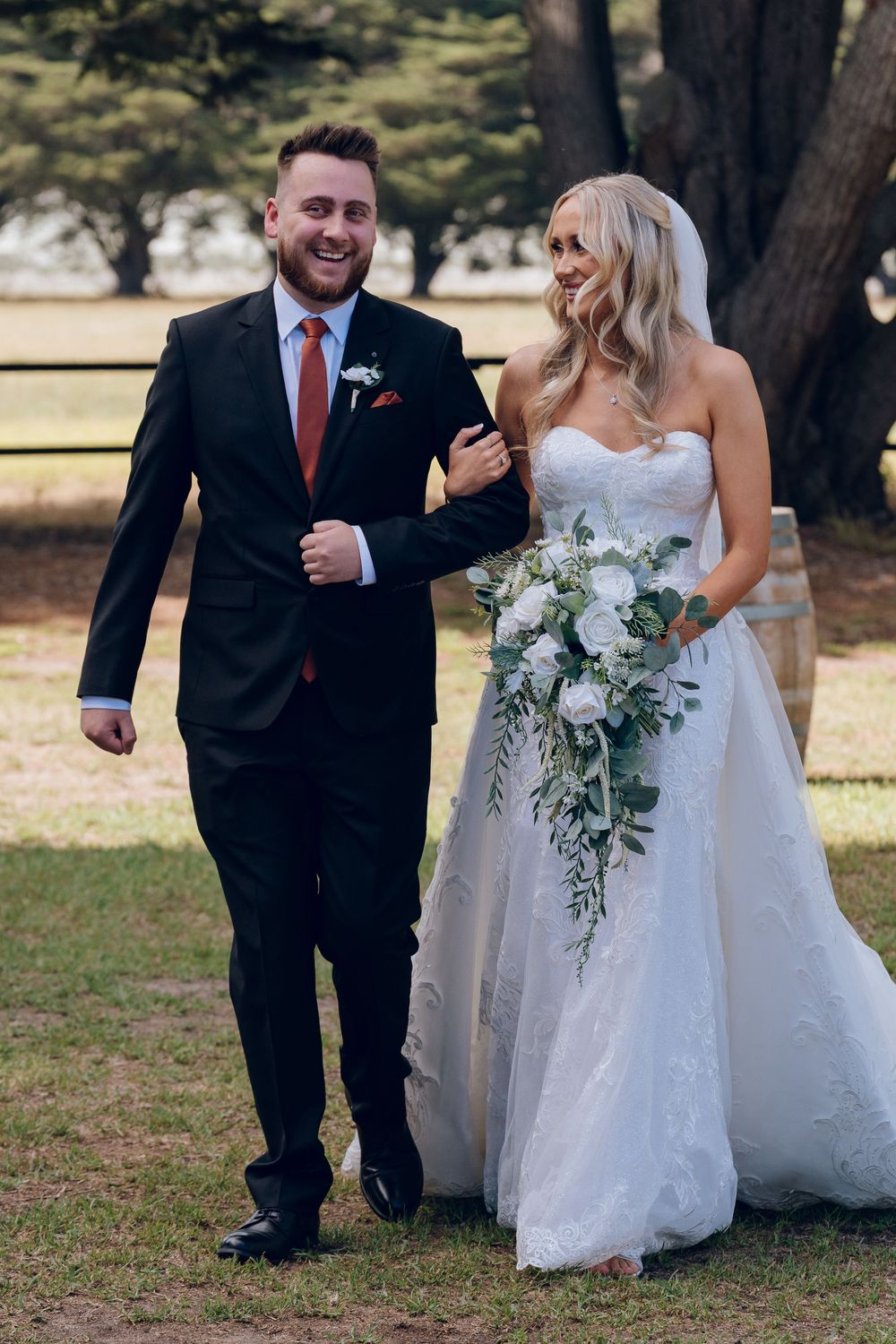 Happy newlyweds walking together outdoors, bride carrying cascading white bouquet.