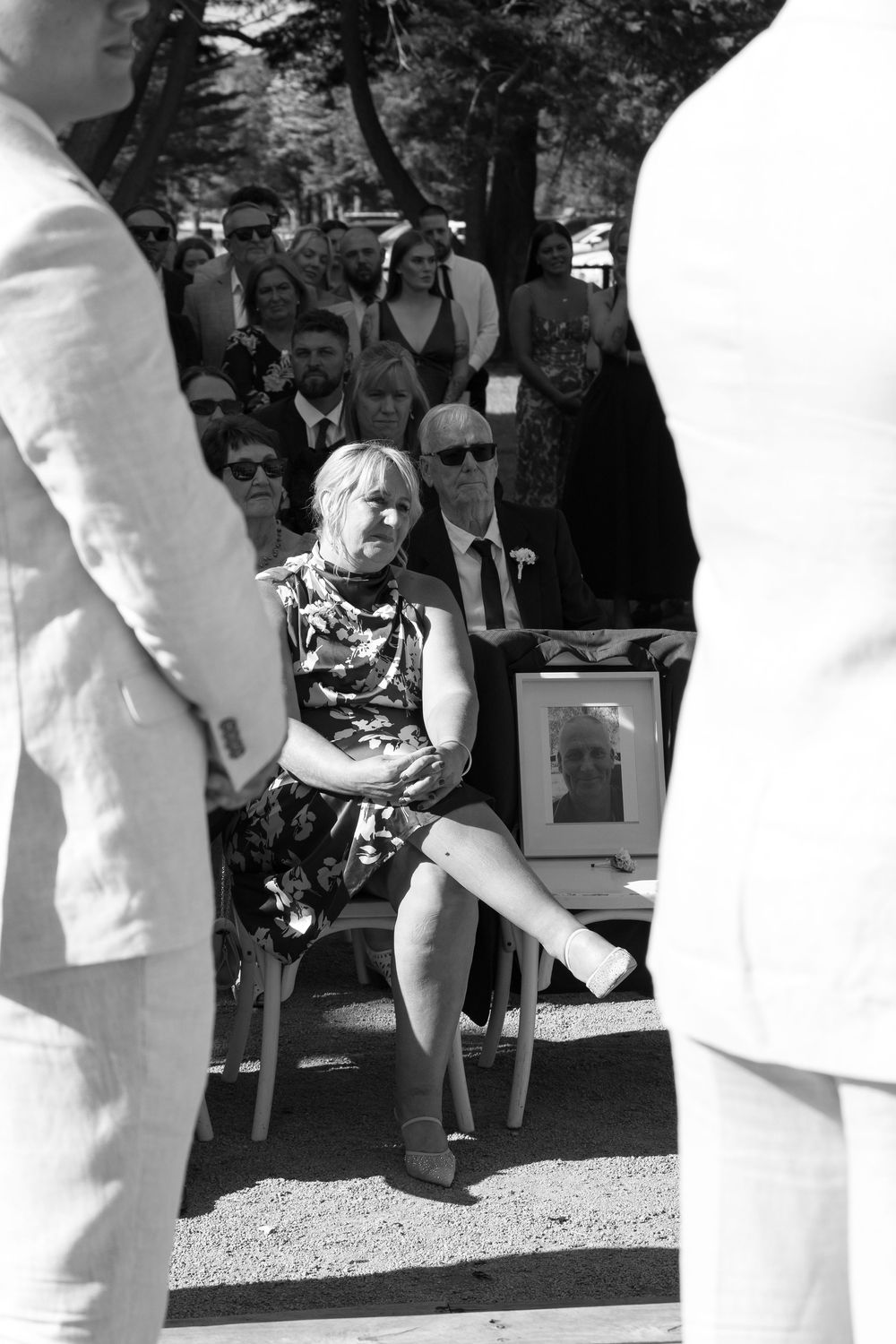 Wedding guests in formal attire gather outdoors for a sunny ceremony.