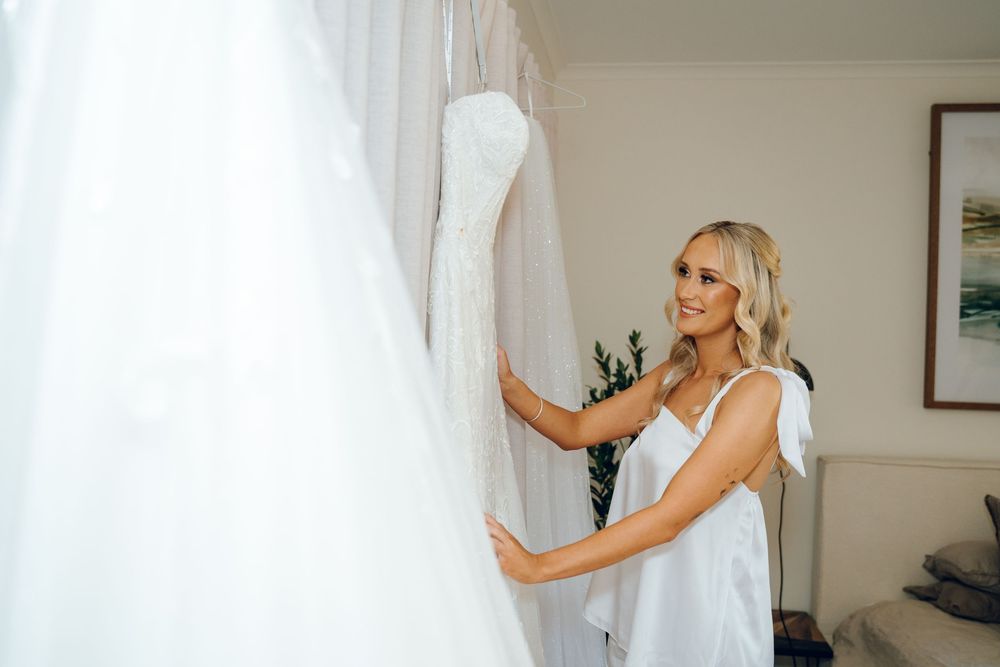 Woman in white dress admires wedding gown hanging before ceremony.
