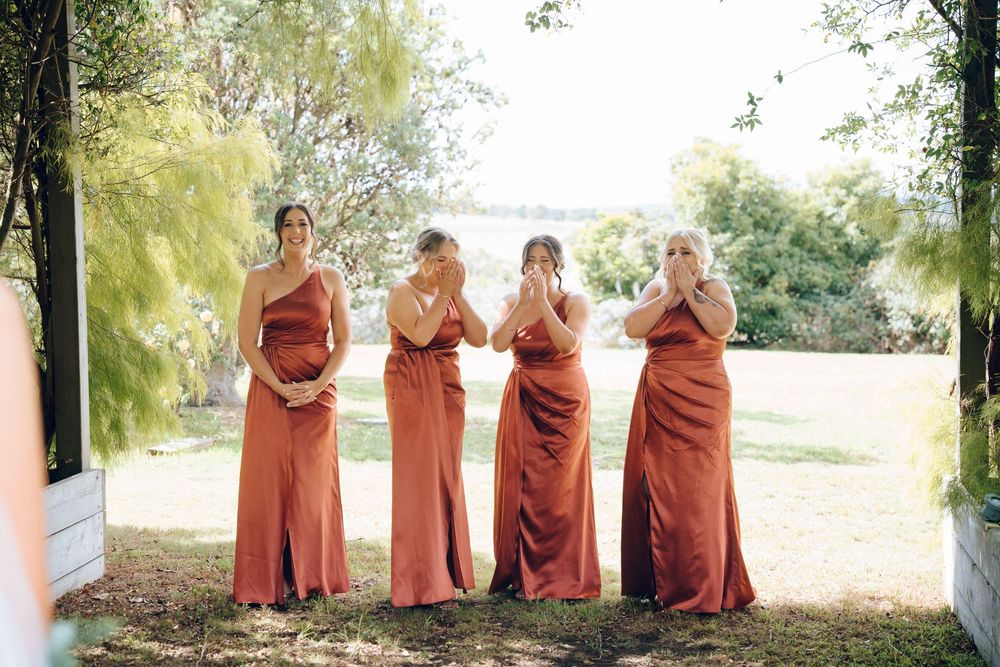 Bridesmaids in rust-colored dresses pose playfully outdoors between white pillars.