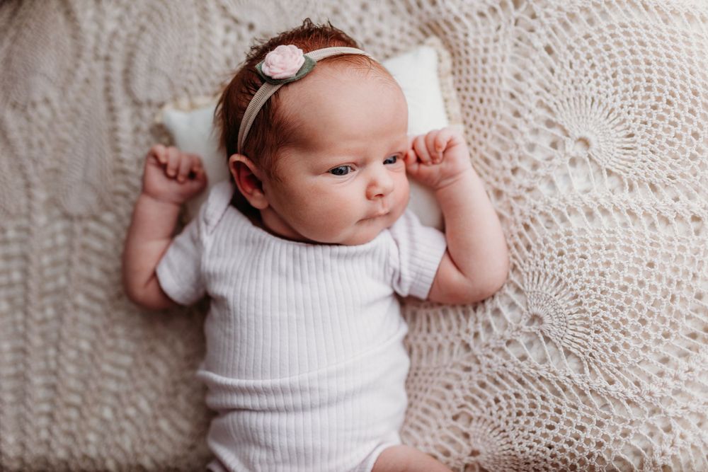 Baby in white onesie lying on cream knitted blanket with hands up near face.