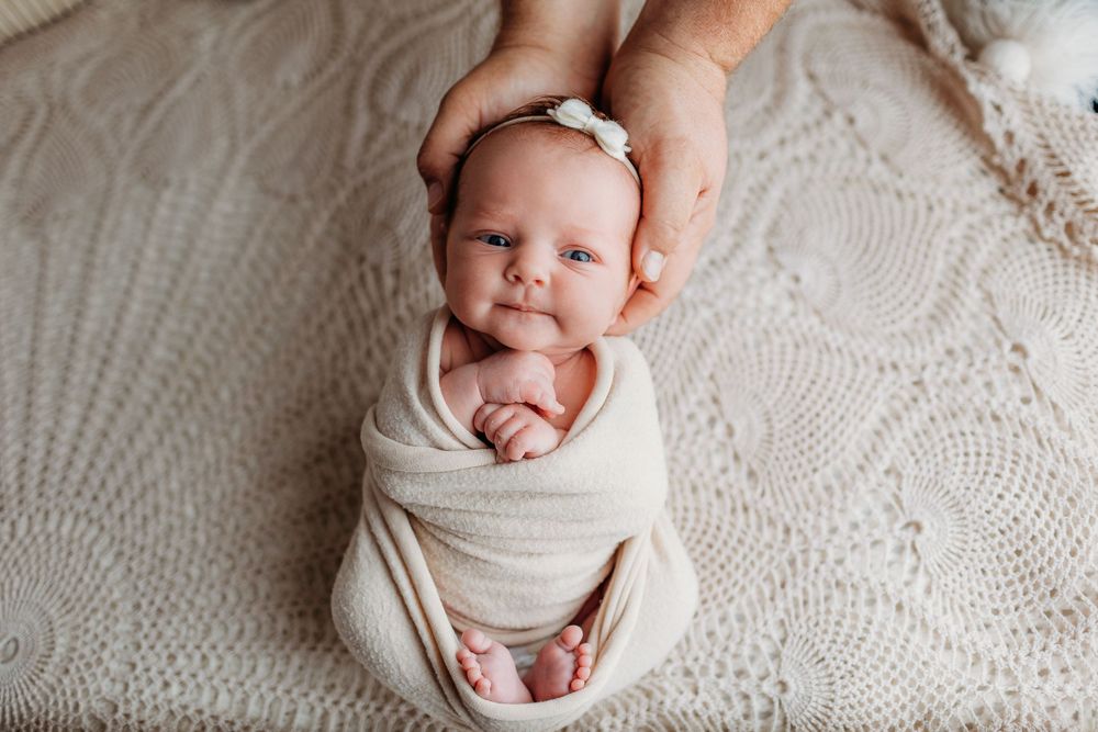 Newborn baby swaddled in white wrap smiling while lying on textured blanket, with hands visible above.