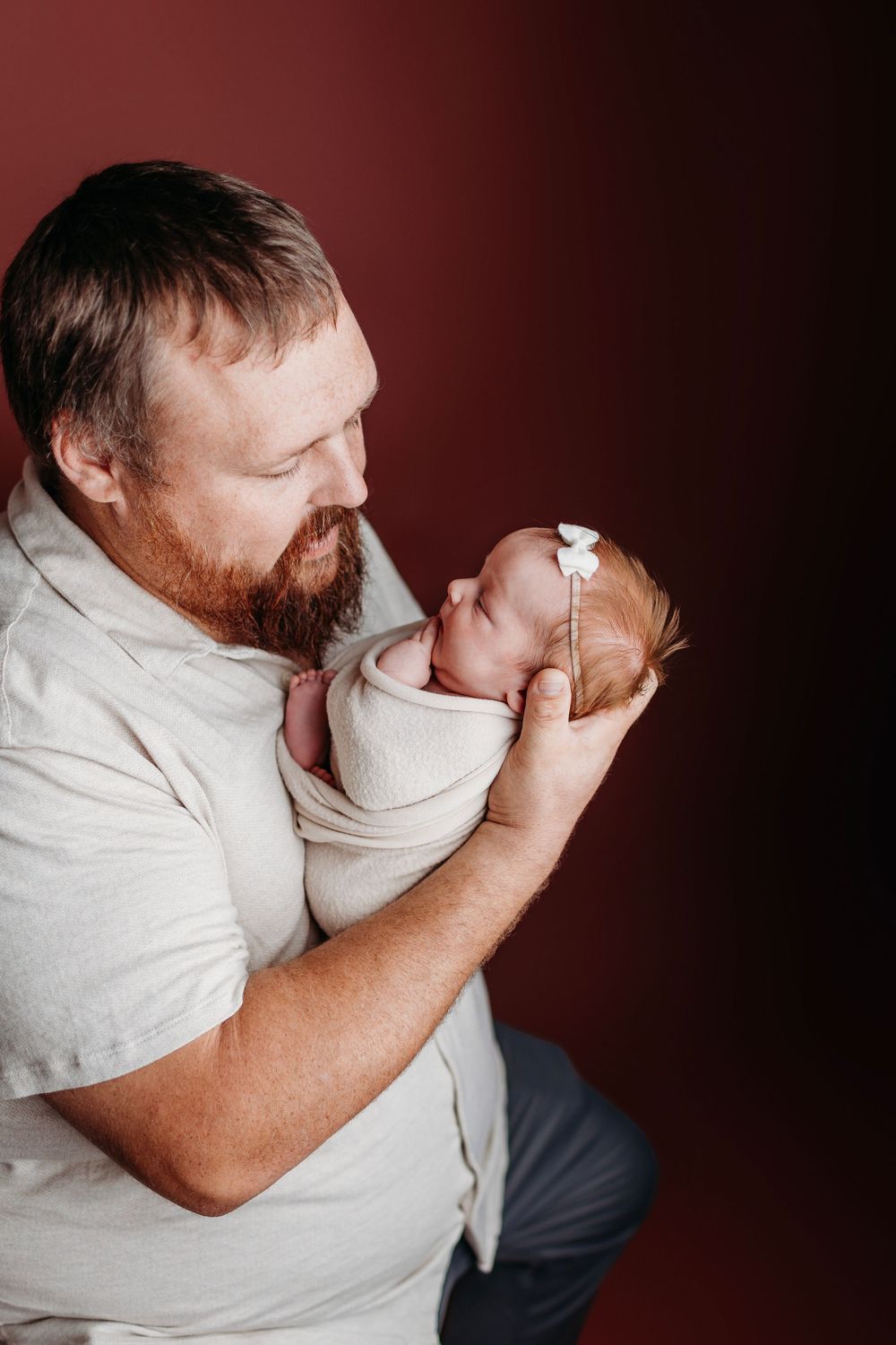 Father in white shirt holds newborn baby against dark red background in tender moment.