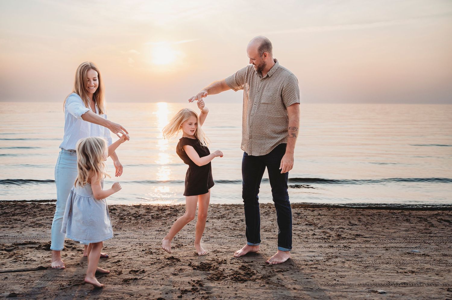 Mississauga family photographer captures mom and dad twirling daughters and smiling on the beach in front of the water during golden hour