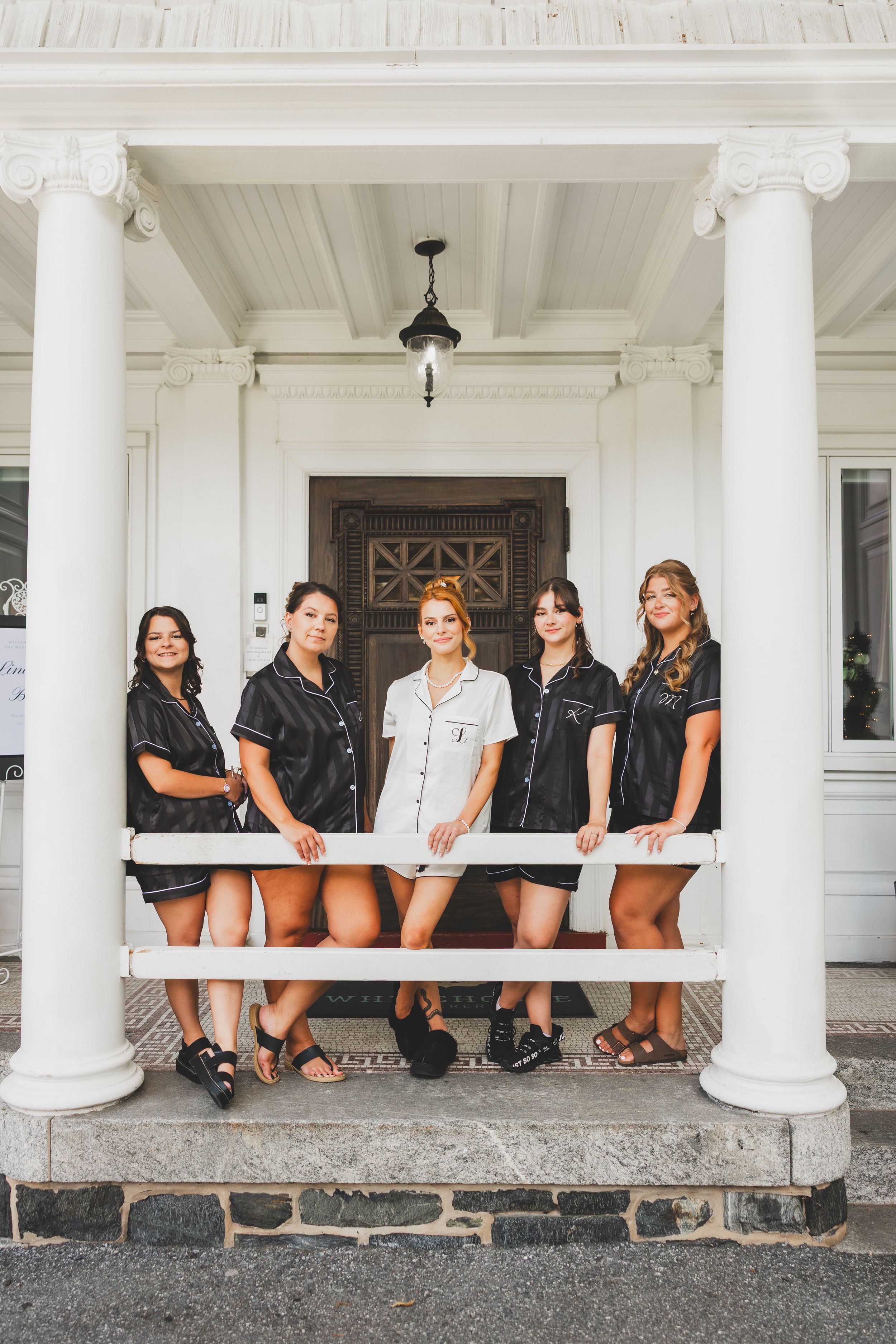 Bride and Bridesmaids stand on a white-columned porch wearing coordinated dress shirts and shorts in a vintage style setting.