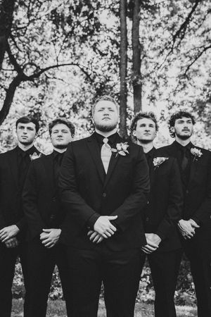 Black and white photo of five groomsmen in formal suits standing outdoors under trees for wedding portraits.