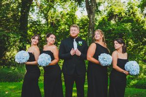 Elegant wedding party wearing black formal attire holds light blue hydrangea bouquets in a sunny garden setting. Groom and bridesmaid photo