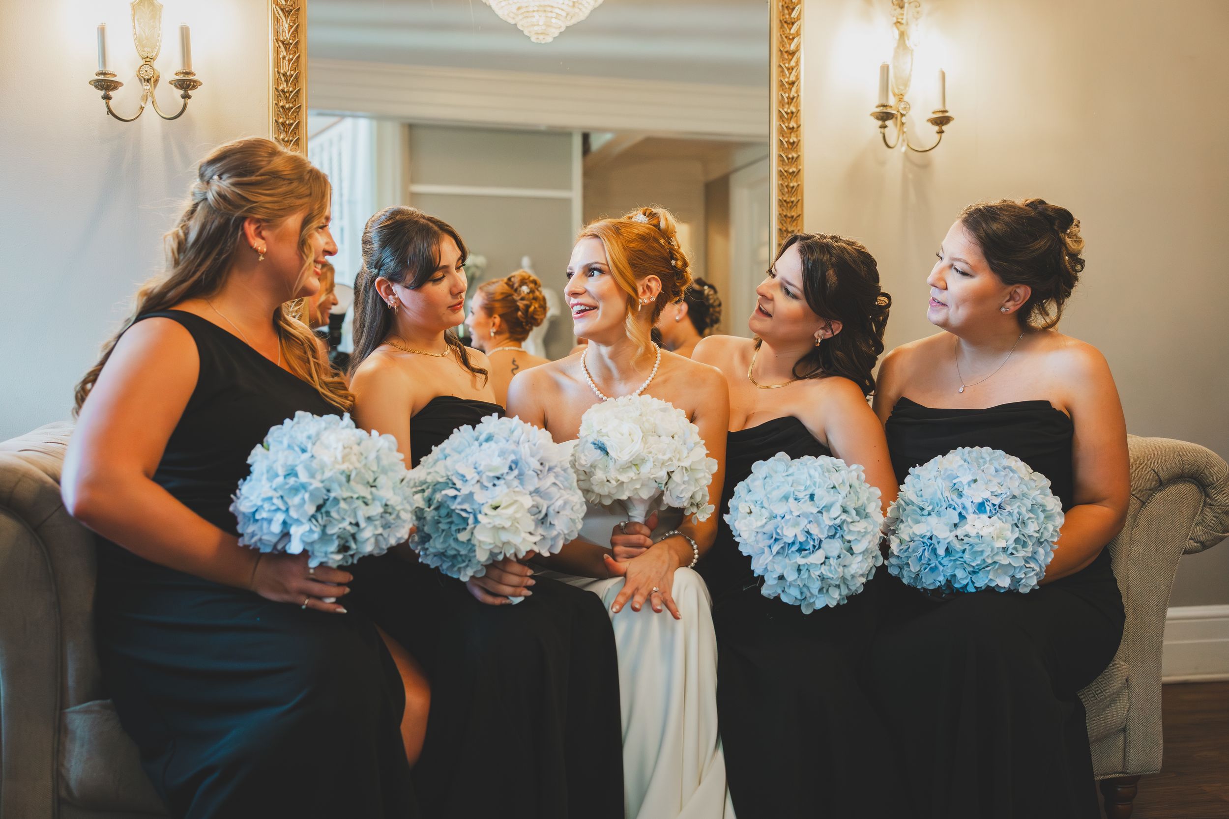 Bridesmaids in navy blue dresses hold pale blue hydrangea bouquets while sharing joyful moments together.