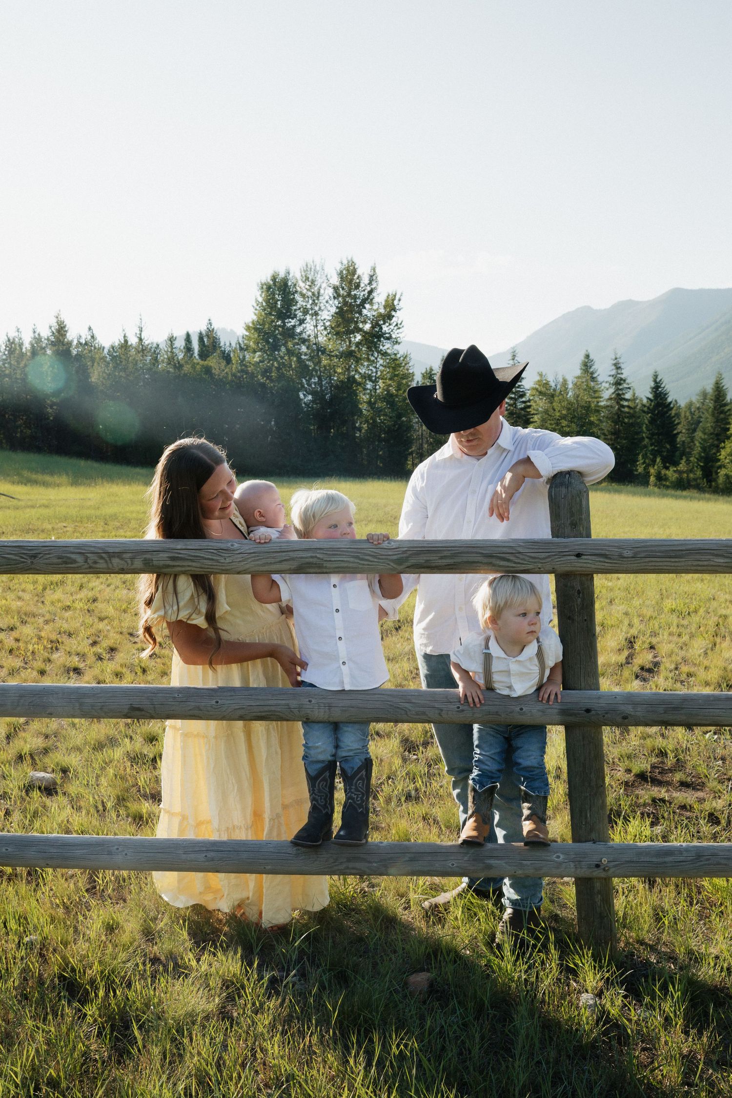 Family wearing coordinated outfits stands together by rustic wooden fence in sunny mountain meadow at sunset.