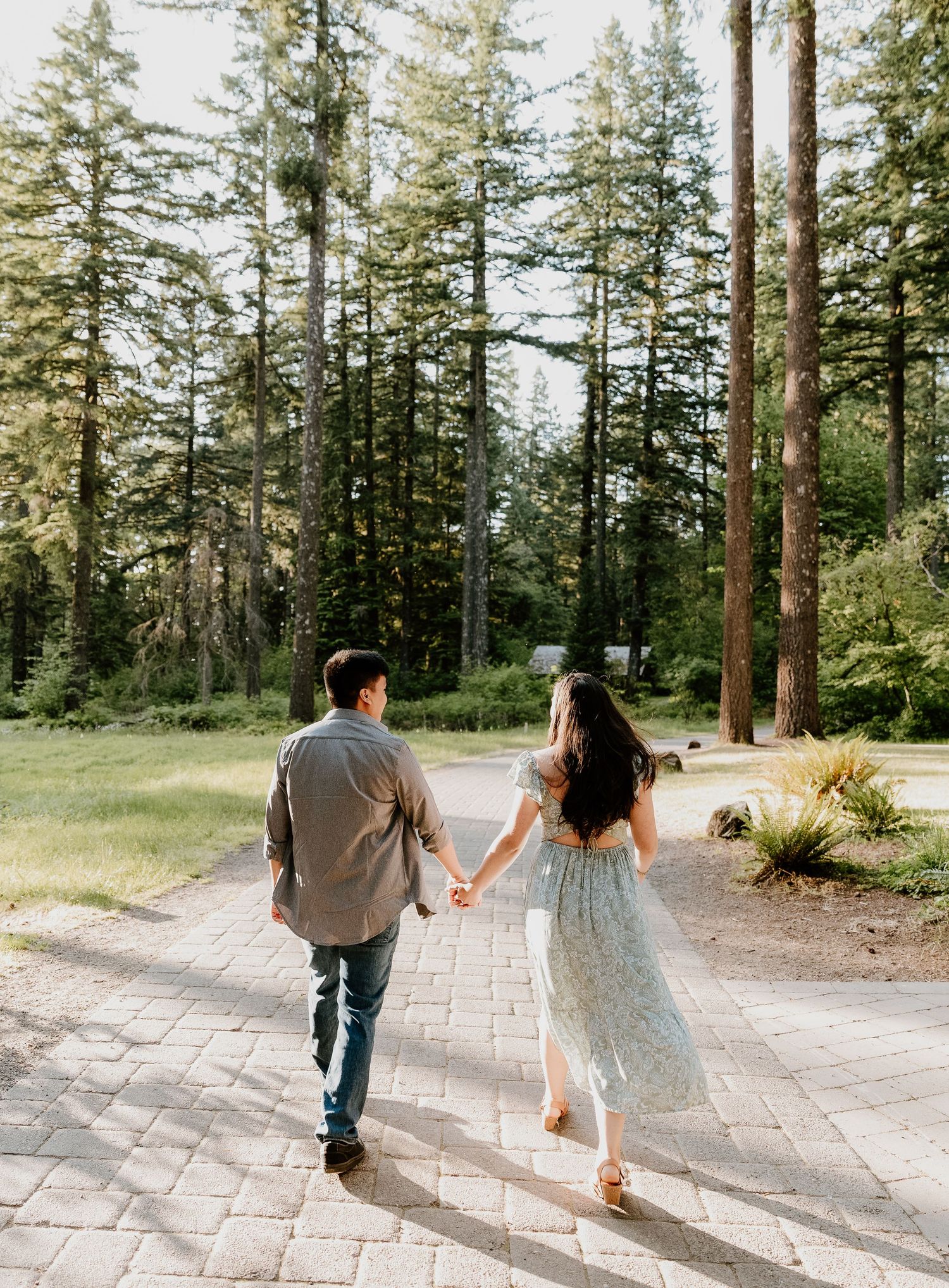 A couple playfully walks down a stone path through a pine forest, holding drinks and creating a sequence of romantic movements.