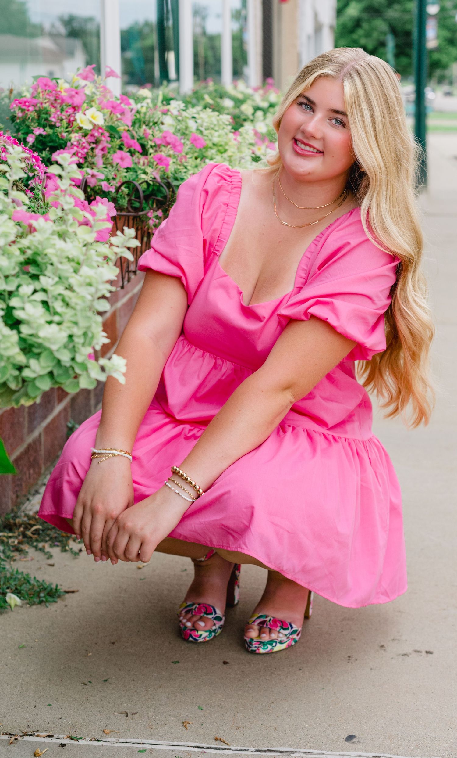 A woman in a bright pink ruffled dress crouches down while posing near flowering bushes and plants.