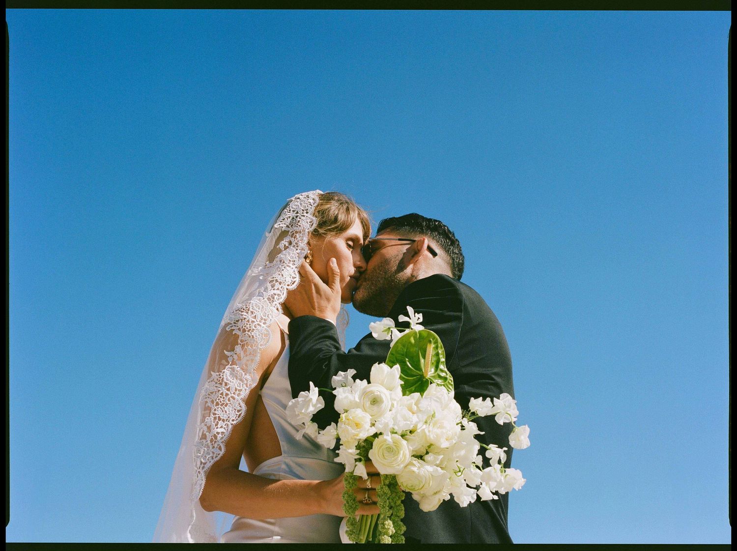 A couple in wedding attire kiss against a bright blue sky while holding a white floral bouquet.