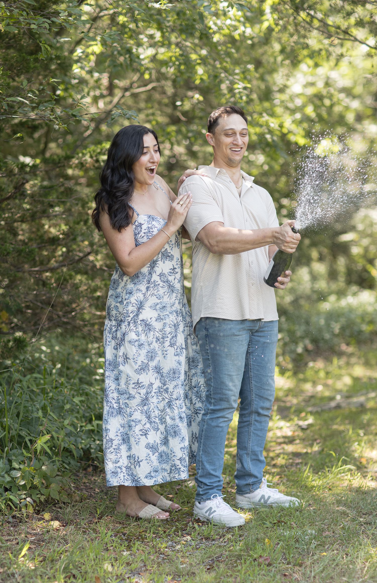 A couple celebrates with champagne outdoors as spray flies from the bottle in a sun-dappled forest setting.