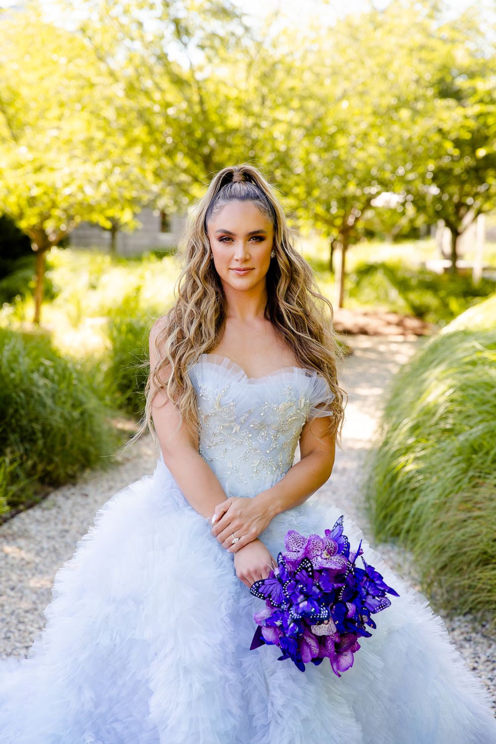 Bride in lace wedding gown holds vibrant purple orchid bouquet while posing in sunny outdoor garden setting.