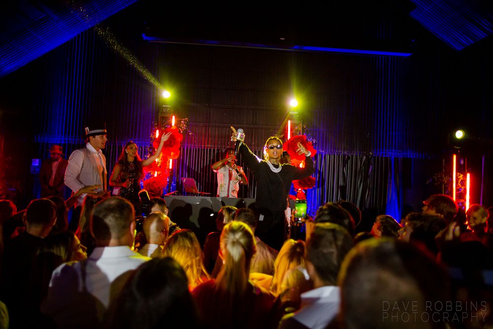 A live band performs on stage at a concert venue with dramatic blue and red lighting while a crowd watches.