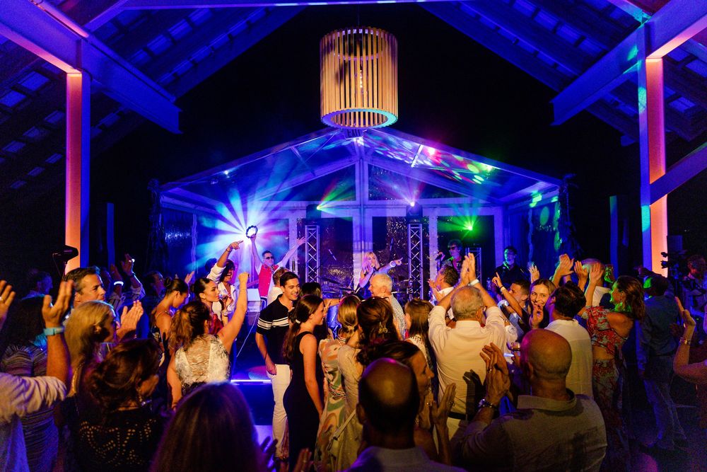 Wedding guests dance and celebrate under purple and blue lighting in an outdoor tent venue.