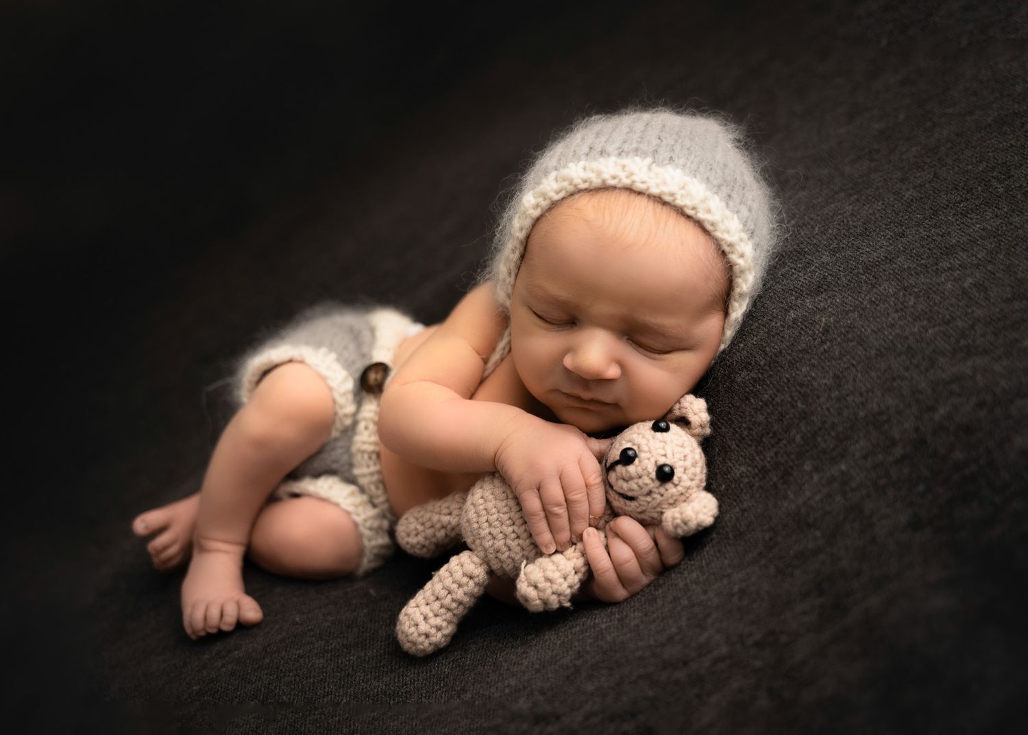 Newborn baby sleeping peacefully on dark background with soft knit teddy bear companion at gently posed newborn session in Arlington Heights