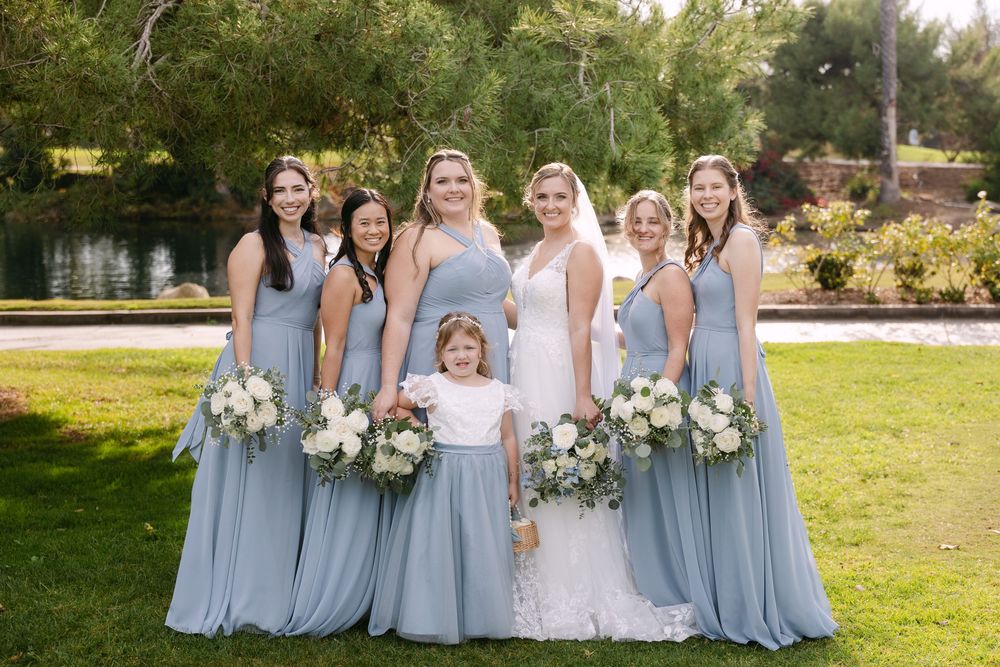 A group in light blue bridesmaid dresses and white wedding gown pose together in a sunny garden setting.