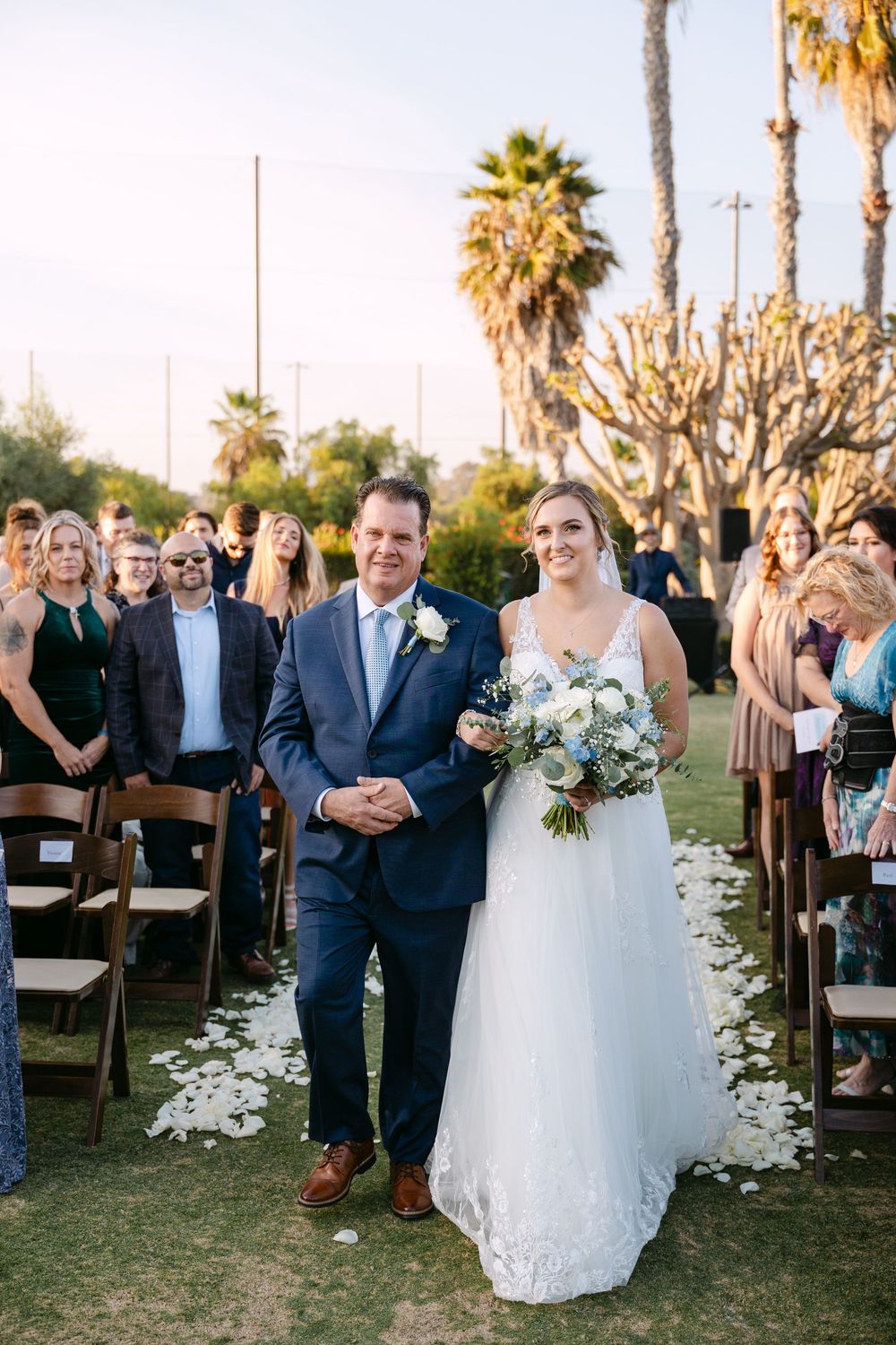 Outdoor wedding ceremony setup with palm trees and scattered white flower petals along the aisle at sunset.
