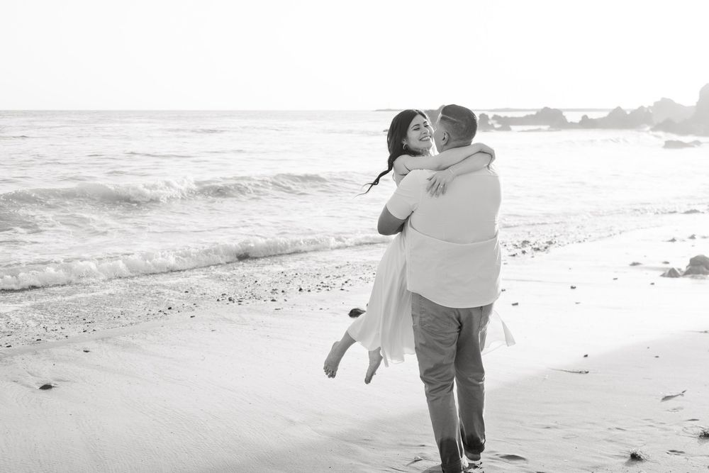 A couple embraces while walking along a misty beach during sunset, creating a romantic silhouette against the ocean.