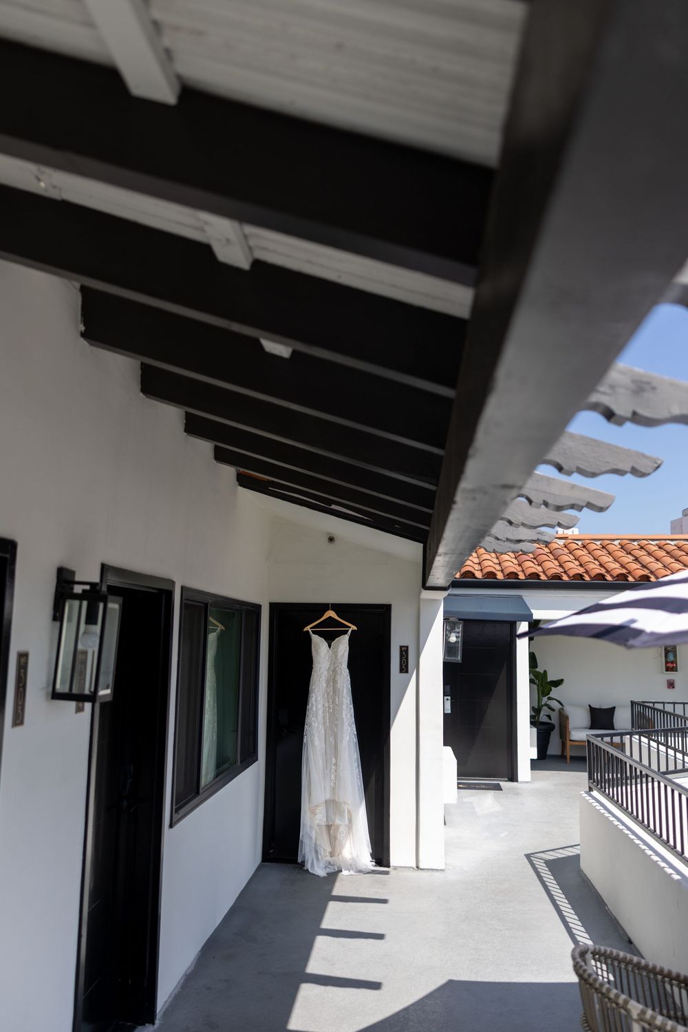 A white wedding dress hangs on a covered outdoor patio with Spanish-style architecture and terracotta roof tiles.