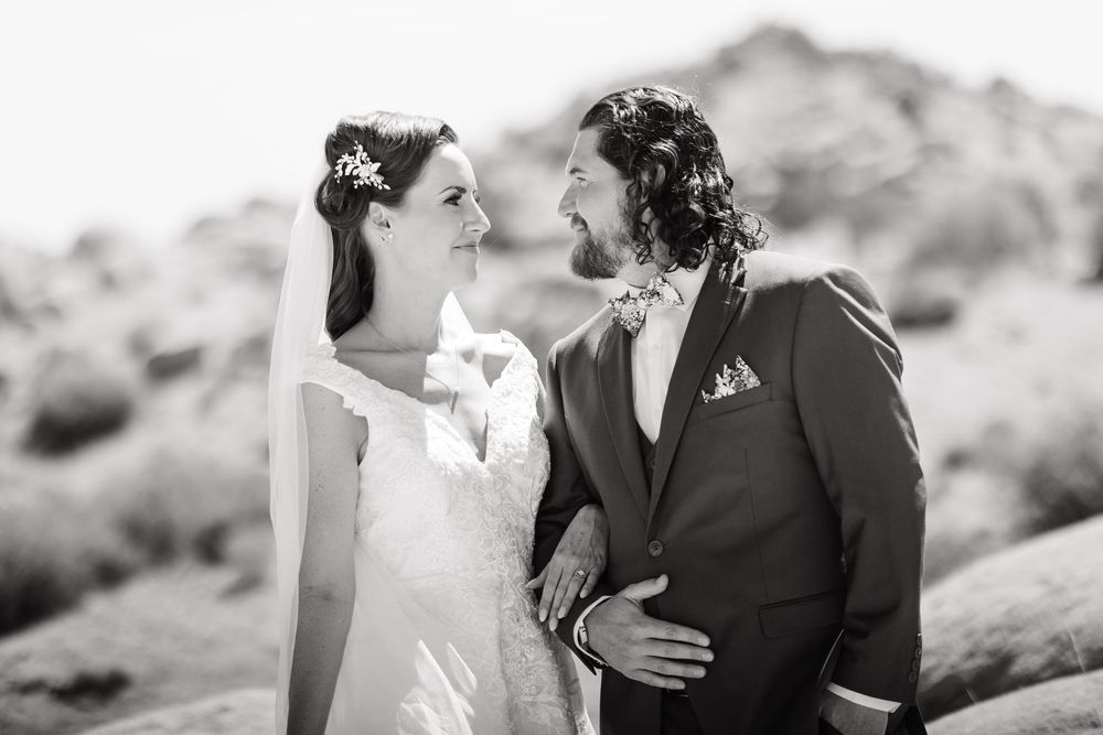 A black and white wedding portrait captures a romantic moment between newlyweds against a mountainous desert backdrop.