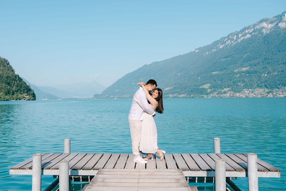 A couple embraces while standing on a wooden pier overlooking a scenic mountain lake on a sunny day.