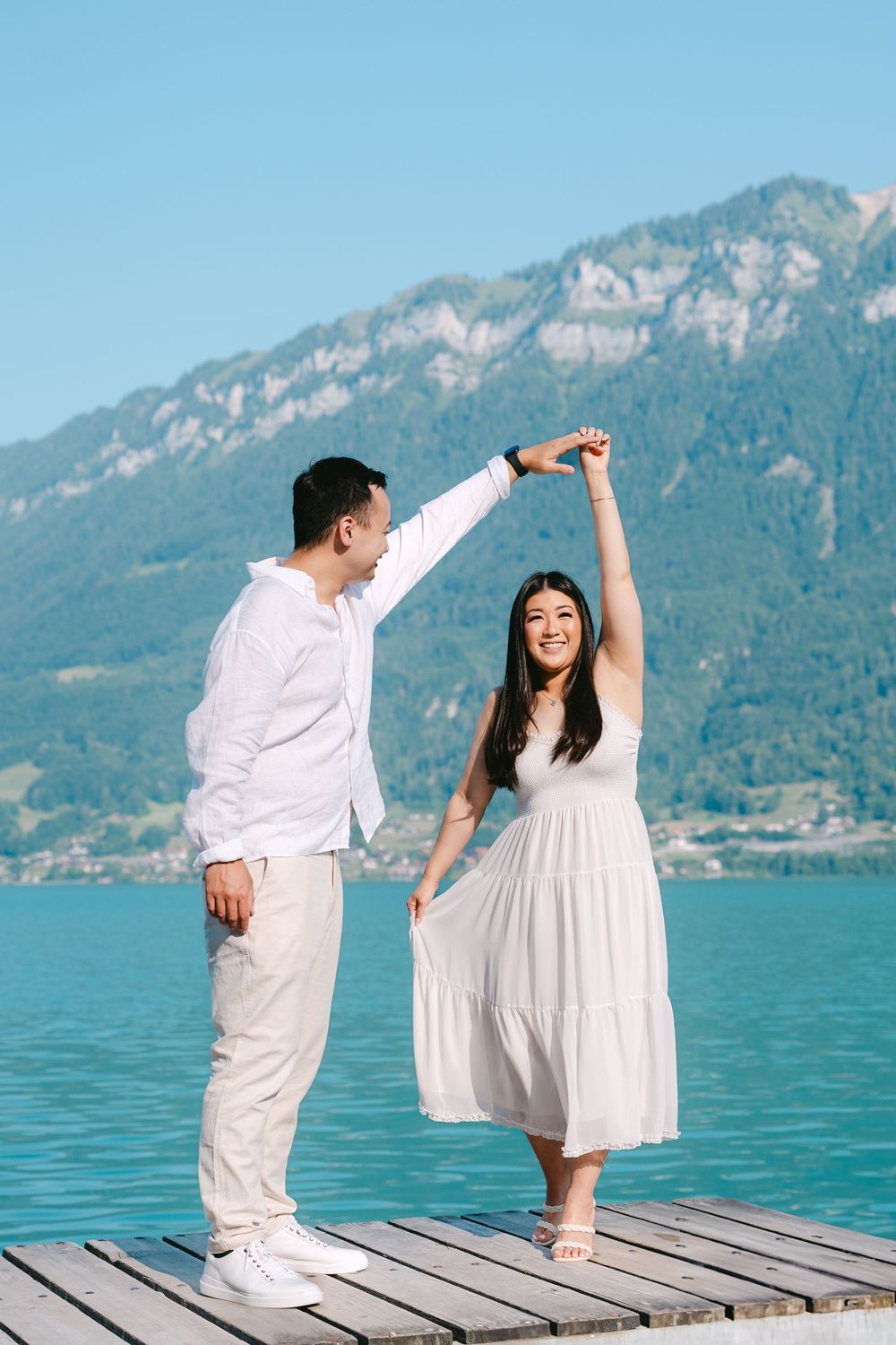 A couple in white outfits dancing playfully on a wooden pier with turquoise lake and mountains in the background.