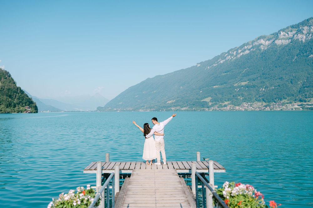 A person in white stands at the end of a wooden pier overlooking a scenic mountain lake with turquoise waters.