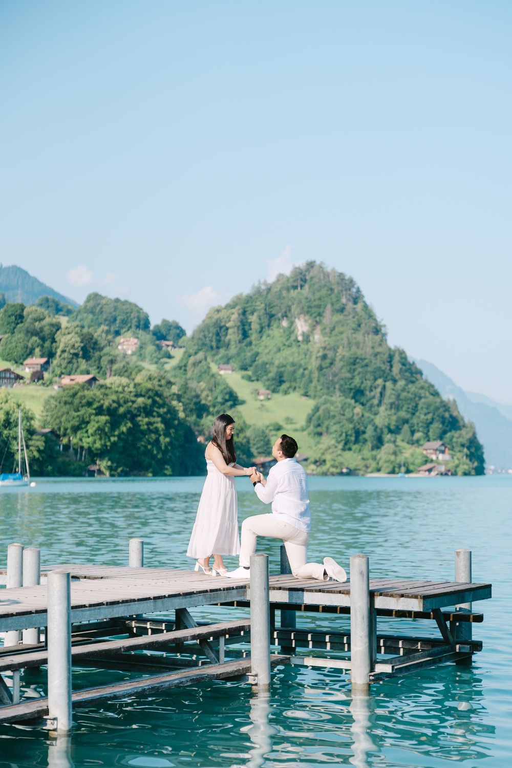A couple in white outfits sits on a wooden dock overlooking a scenic mountain lake.