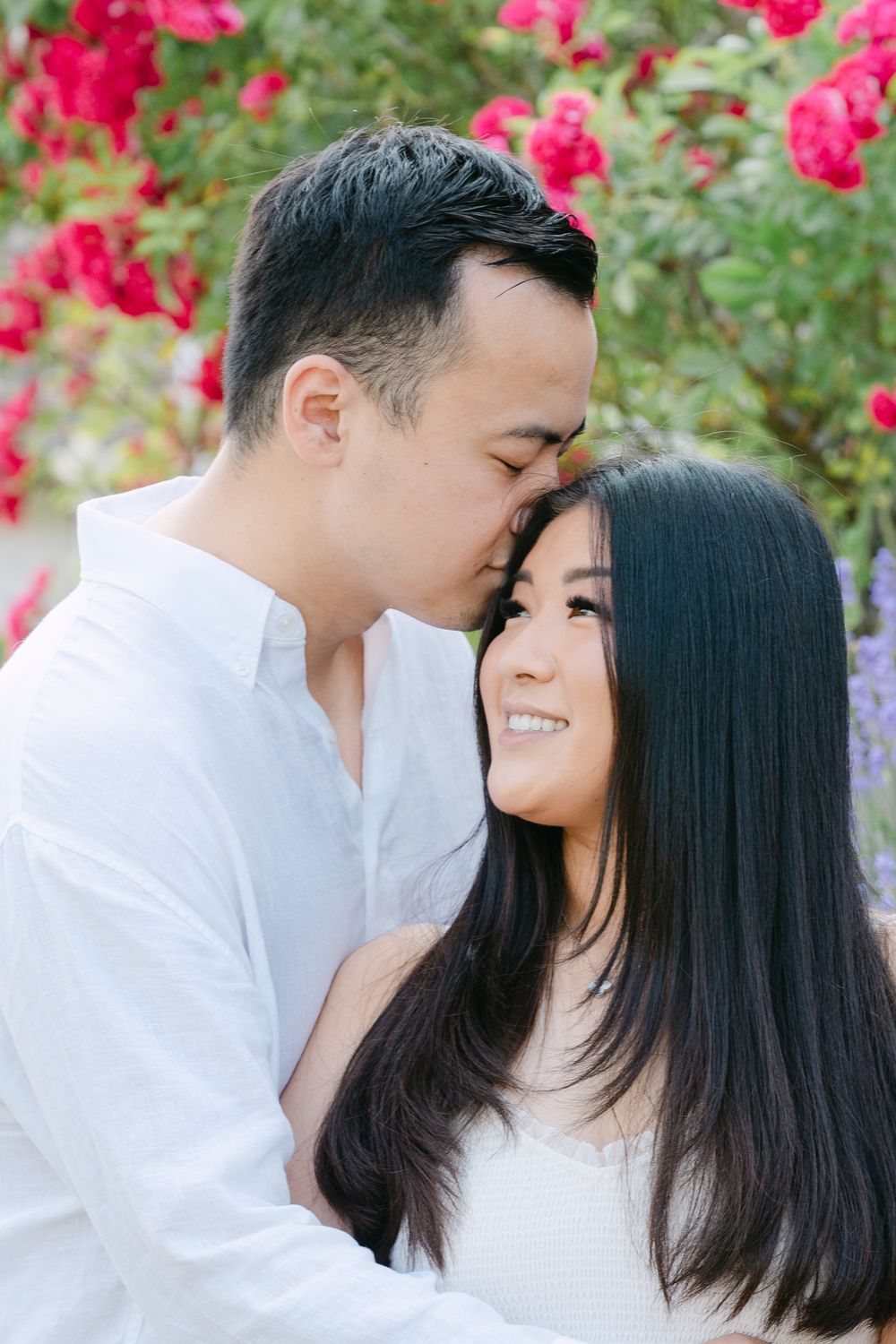 A romantic couple embracing in front of vibrant pink roses and purple flowers in a garden setting.