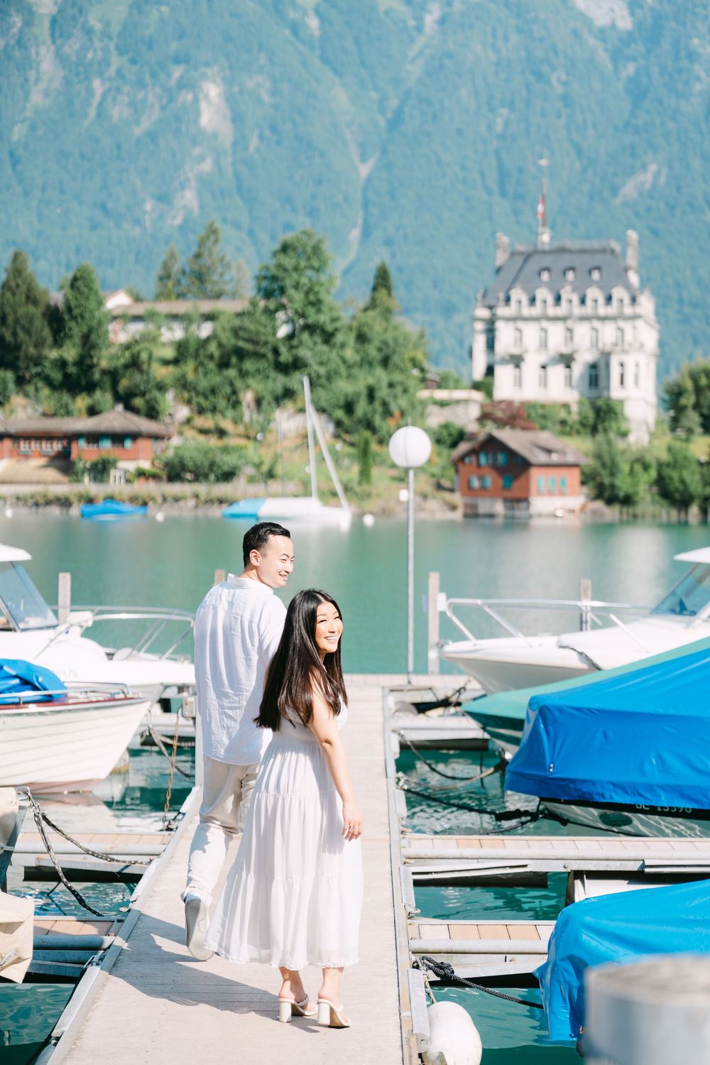 A couple walks along a marina dock with boats and a grand mansion in the mountain backdrop.
