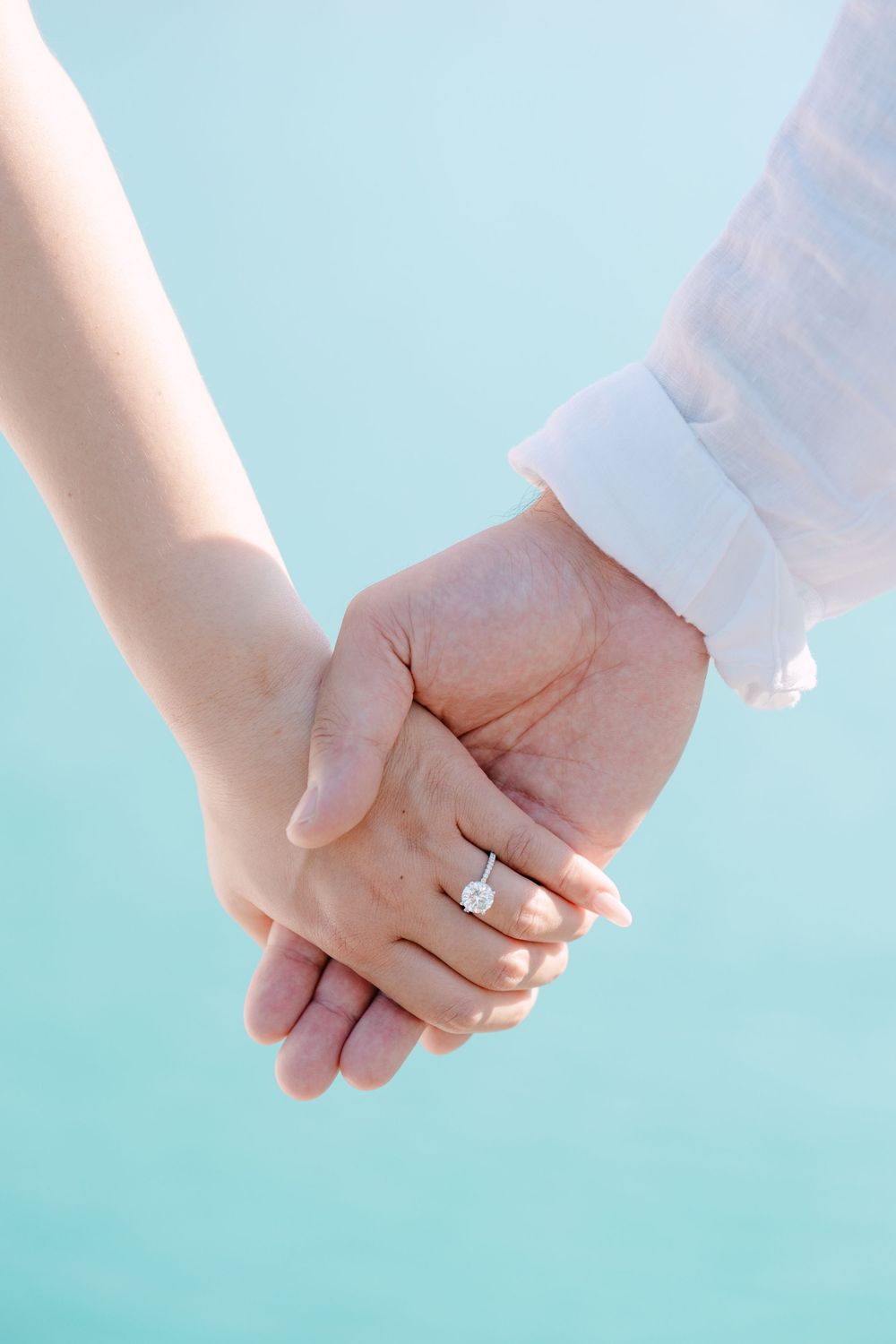 Two hands clasped together with a wedding ring visible against a soft turquoise background.