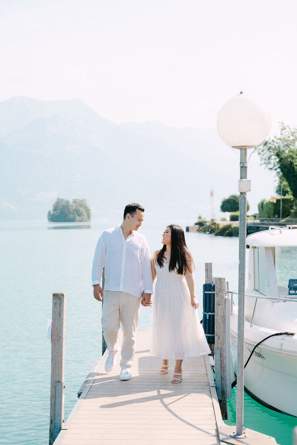 Two people stroll hand in hand on a lakeside dock with mountains and boats in the background.