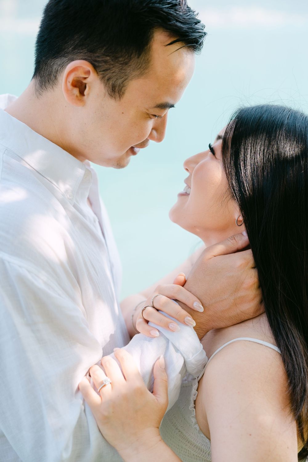 An intimate portrait of a couple in white embracing and smiling against a soft blue background.