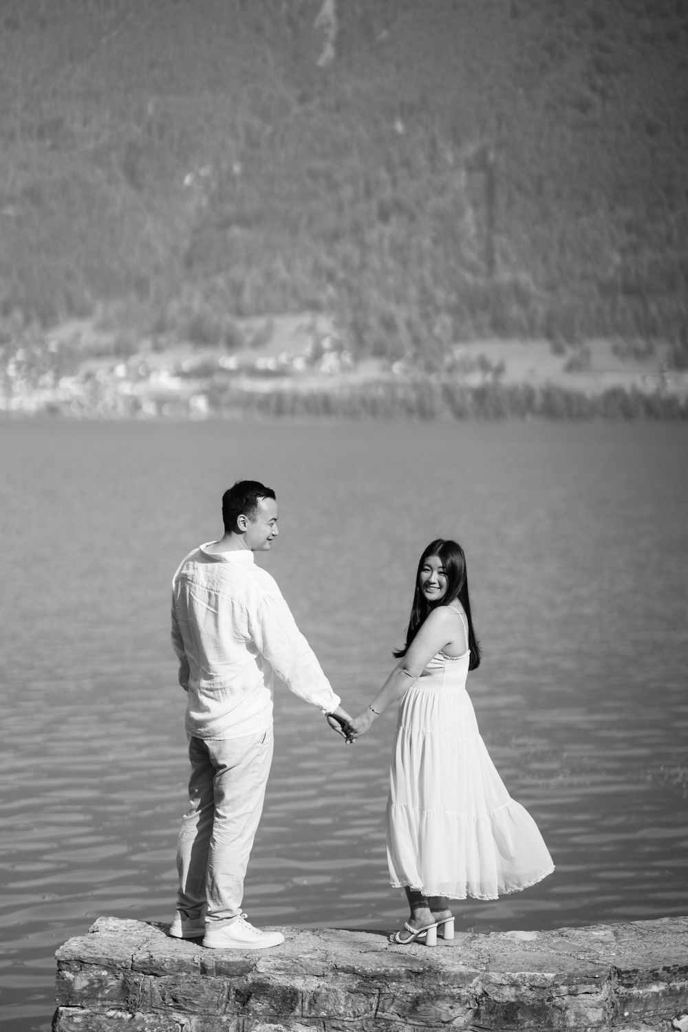 A black and white photo of two people standing on a rock by a serene mountain lake.
