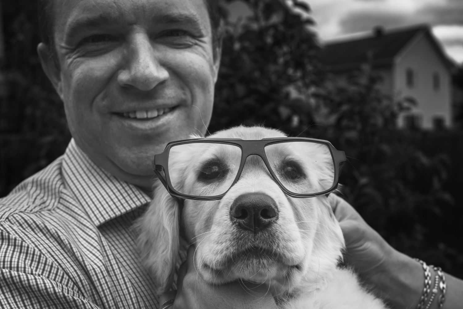 Black and white image of a man holding a puppy that is wearing his human glasses and has a funny look on their face.