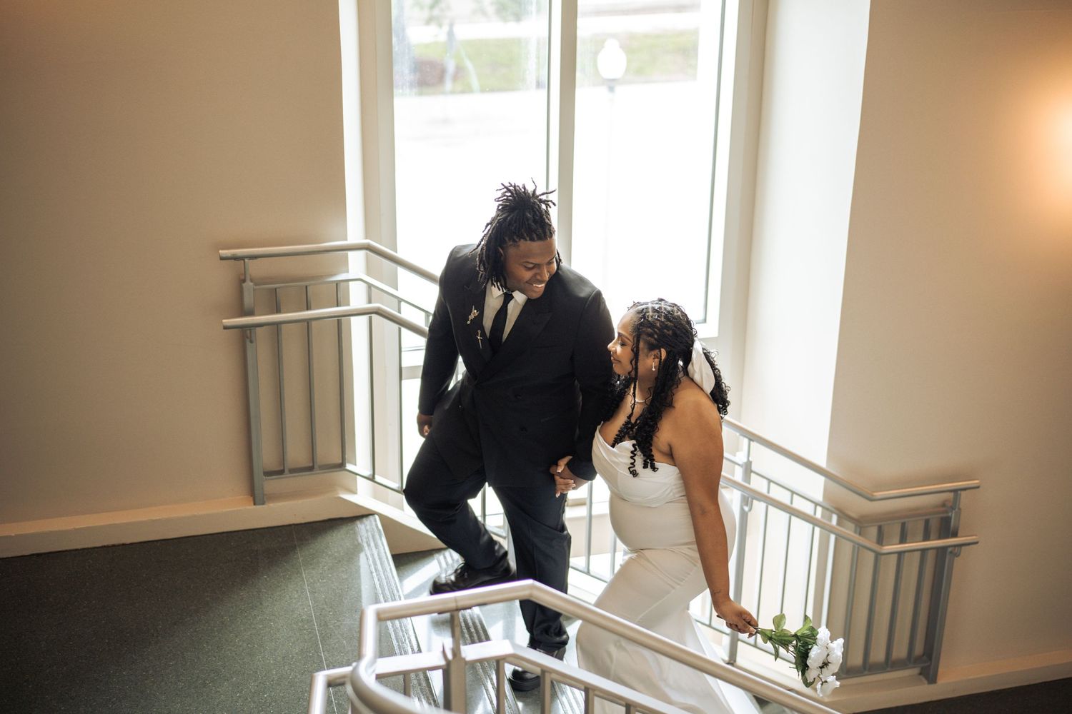 A couple in formal wedding attire descends an elegantly lit interior staircase together.
