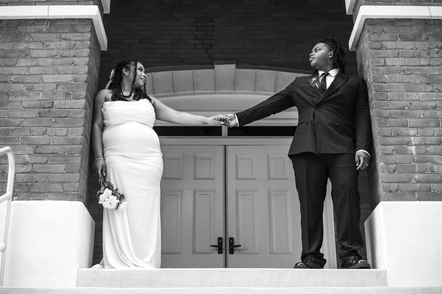 Wedding party poses in joyful, candid moment against brick wall with white doorway during outdoor celebration.